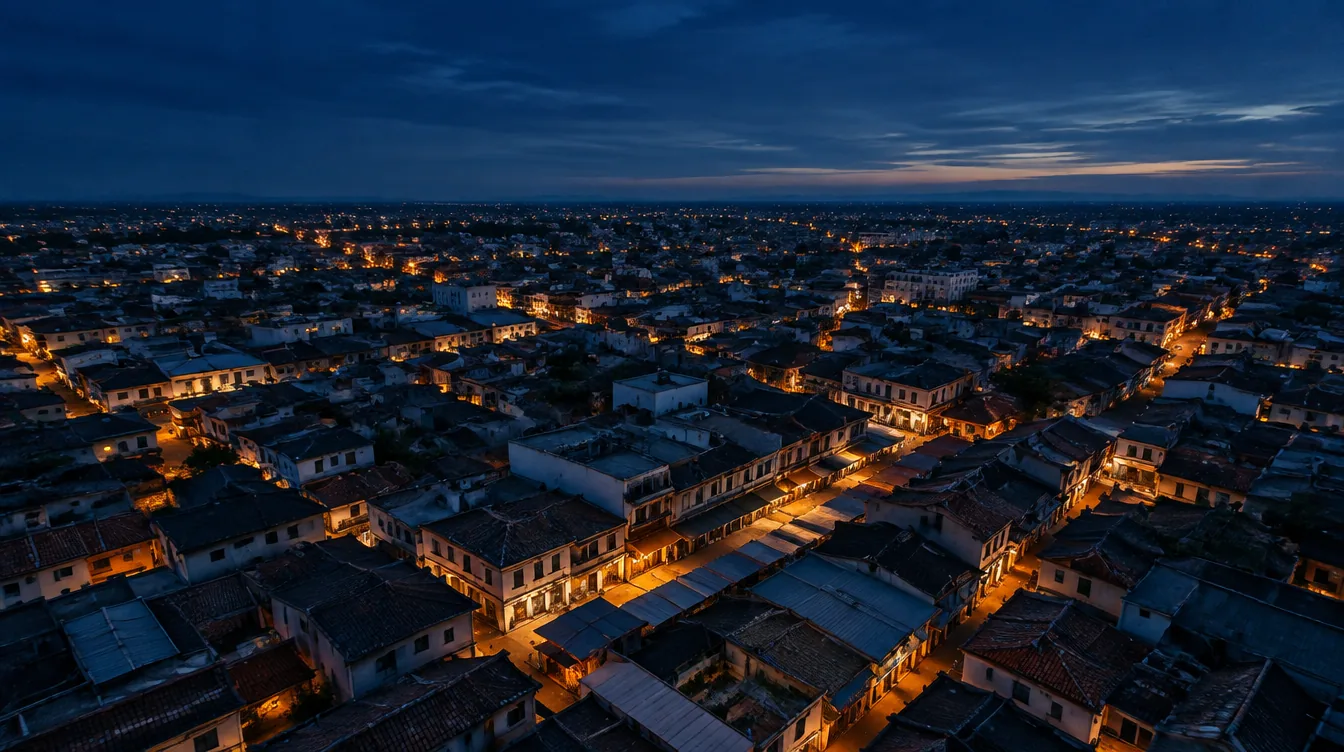 Vue aérienne des toits d'une ville dense à l'heure bleue, les lampadaires ambre s'allumant le long des rues jusqu'à l'horizon.